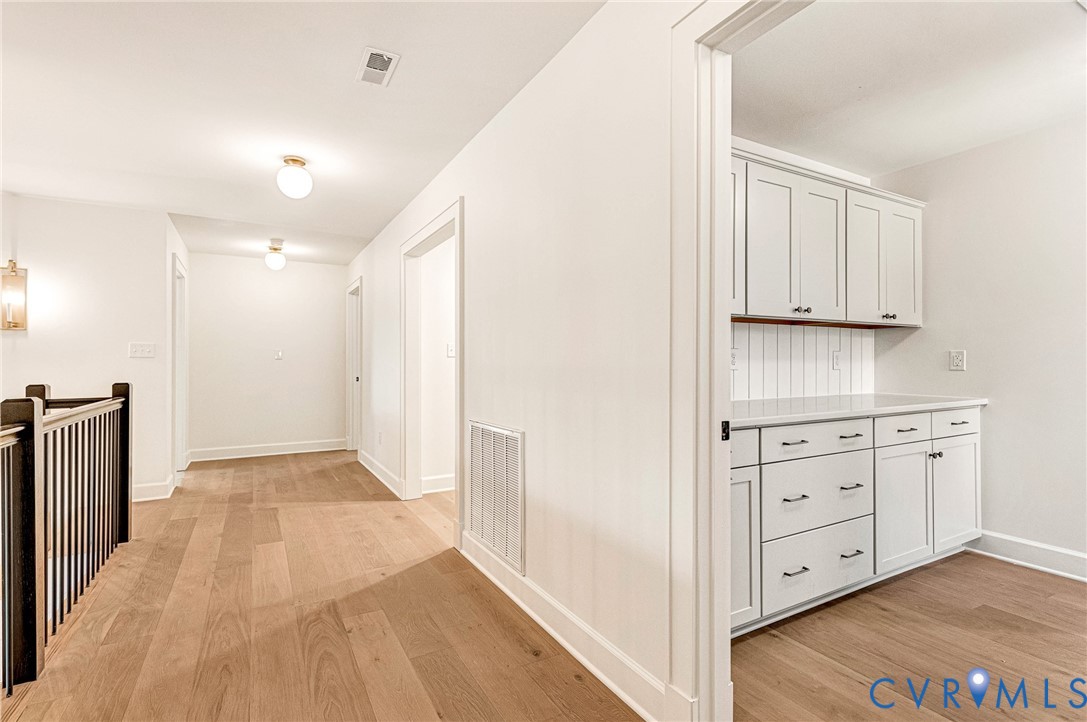 Interior hallway featuring wood-finish flooring, white cabinetry with vertical plank backsplash, and a staircase with a dark-finish railing