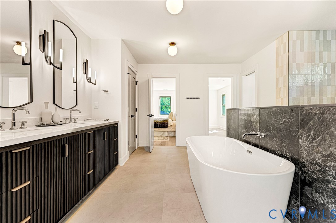 Dual-sink vanity with fluted cabinetry and light-toned countertops, freestanding soaking tub, dark stone surround, wall-mounted faucet, and arched-frame mirrors