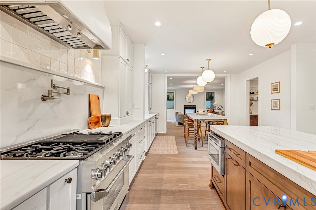 Kitchen featuring white shaker-style cabinetry, solid-surface countertops with continuous backsplash, stainless steel professional-grade range, wall-mounted pot filler, and wood-finish flooring