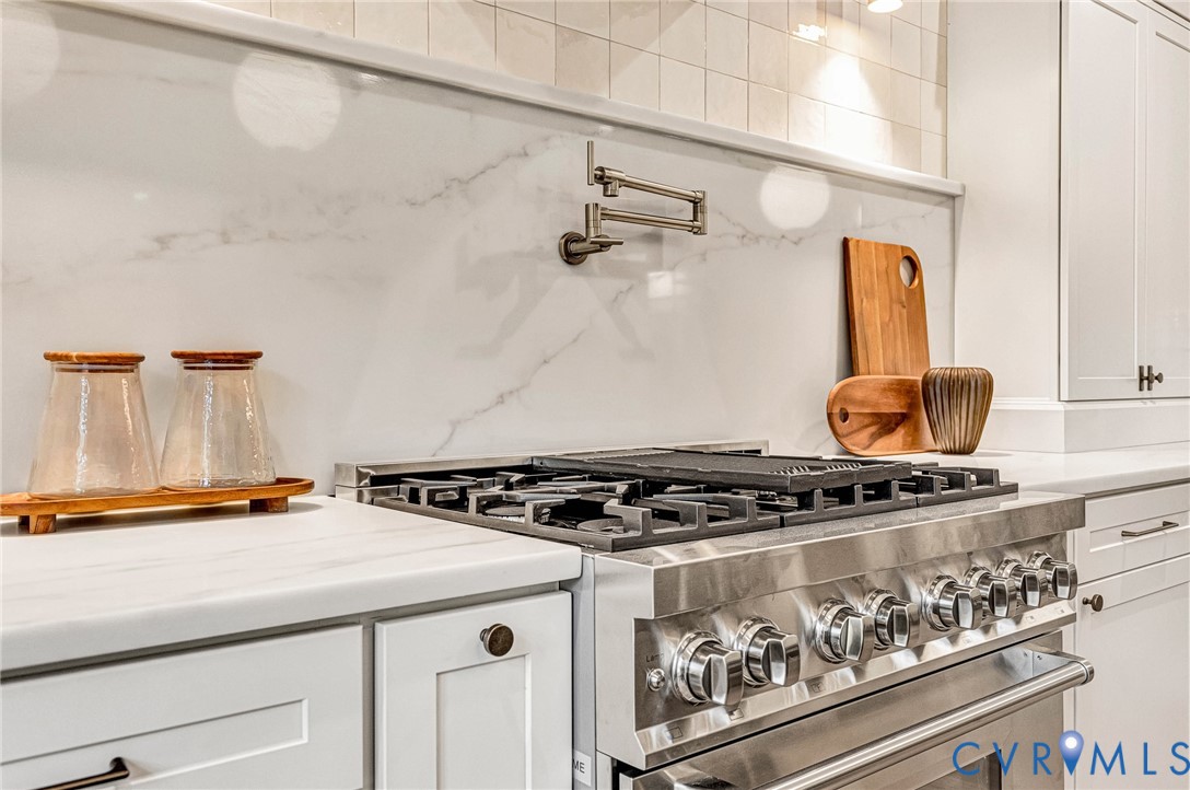 Kitchen featuring a stainless steel gas range with a center griddle, wall-mounted pot filler, solid-surface backsplash, white cabinetry, and light-toned countertops