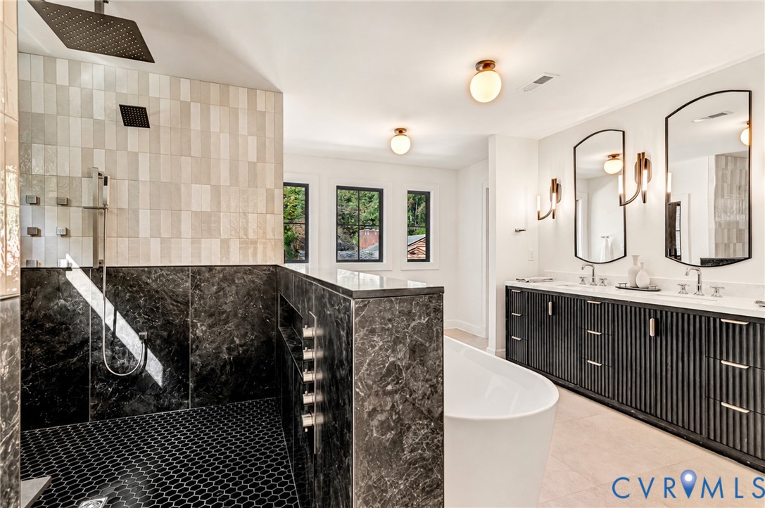 Walk-in shower featuring a rain-style showerhead, dark stone tile wall, and mosaic floor tile; deep soaking tub; dual-sink vanity with fluted cabinetry; arched wall mirrors with integrated lighting