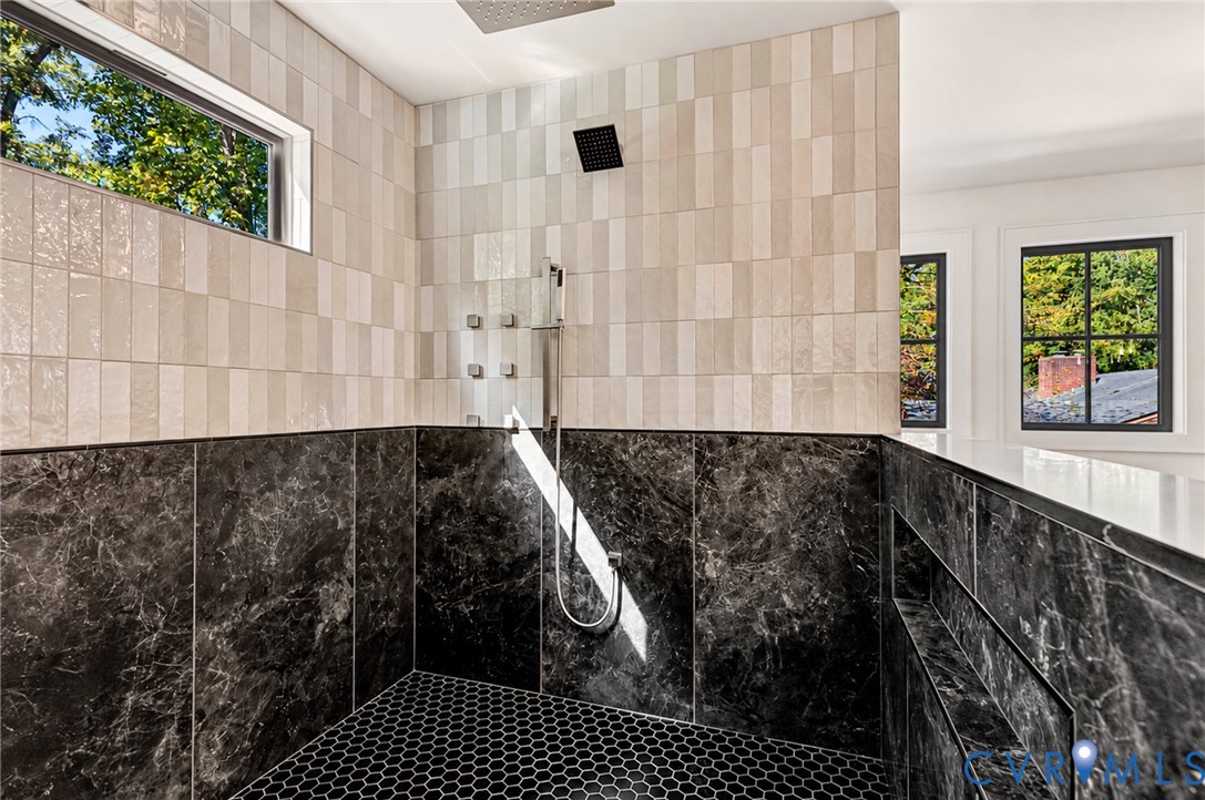 Walk-in shower featuring large-format dark stone wall tiles, cream-toned vertical subway wall tiles, a rainfall showerhead with integrated body jets, and hexagonal floor tiling