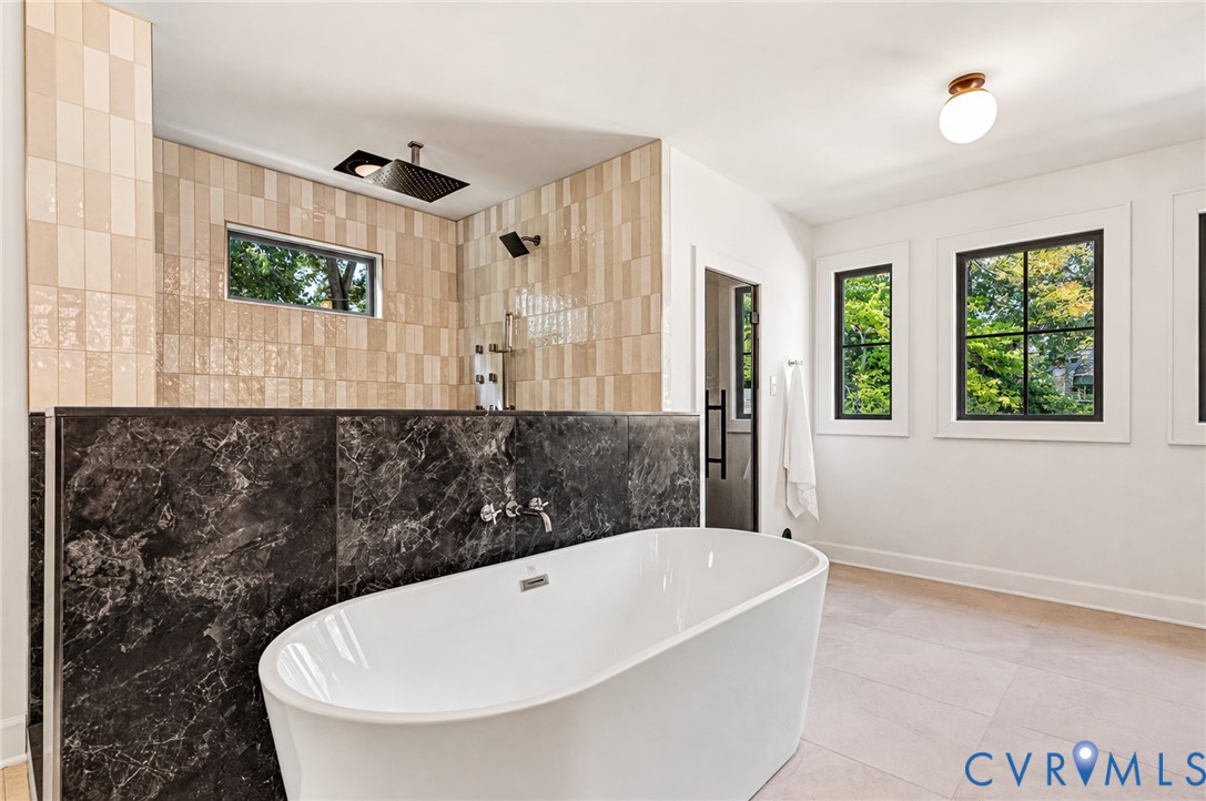 Freestanding soaking tub with wall-mounted plumbing fixtures, dark natural stone half-wall, stacked tile shower surround, and multi-pane windows with dark frames