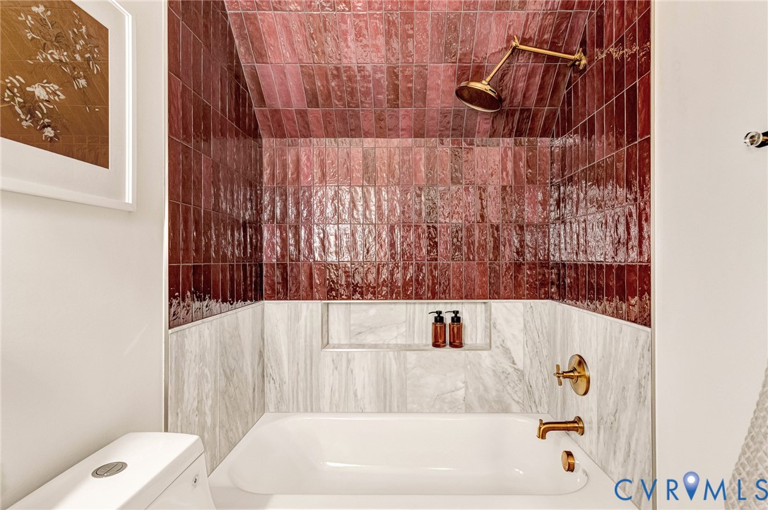 Tub and shower alcove featuring floor-to-ceiling gloss-finish burgundy subway tile, white stone-patterned tile wainscoting, a built-in recessed storage niche, and brushed brass-finish plumbing fixtures