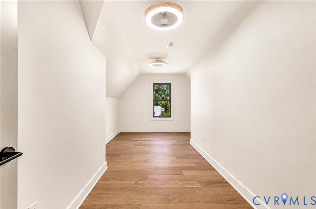 Neutral-toned room featuring wood-finish flooring, sloped ceilings, two circular flush-mount light fixtures, and a single window with dark framing
