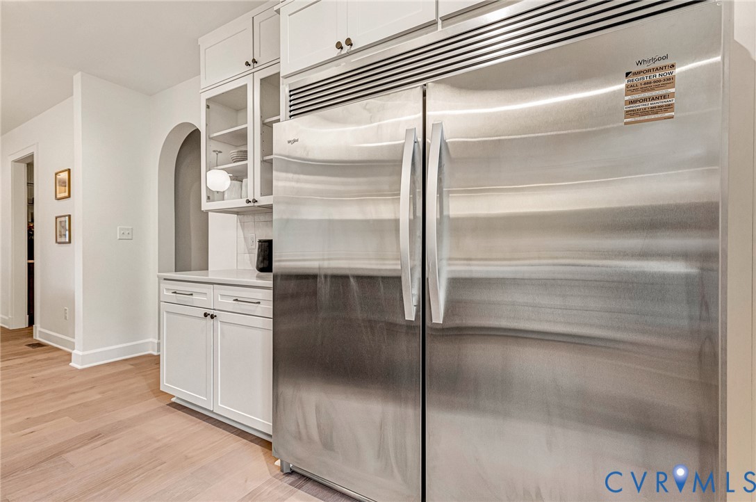Stainless steel side-by-side refrigerator, white shaker-style cabinetry with glass-front uppers, wood-finish flooring, and an arched wall opening