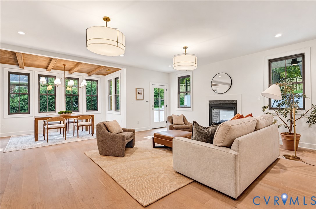 Open-concept interior featuring light wood-finish flooring, exposed wood ceiling beams, a neutral-toned fireplace with a dark surround, and dual drum pendant light fixtures
