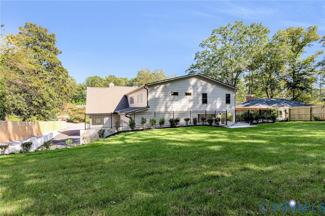 Expansive backyard featuring a manicured lawn, metal perimeter fencing, mature trees, a covered patio structure, and a paved driveway
