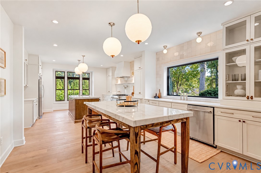 Kitchen featuring an expansive stone-topped island with wood base, white shaker-style cabinetry, recessed and globe pendant lighting, tile backsplash, and light-tone wood-finish flooring Kitchen featuring an expansive stone-topped island with wood base, white shaker-style cabinetry, recessed and globe pendant lighting, tile backsplash, and light-tone wood-finish flooring