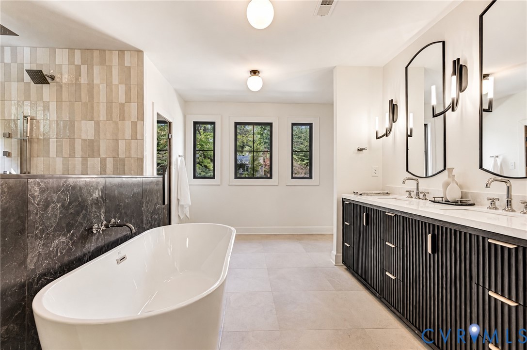 Freestanding soaking tub paired with a stone-accented partition, dual-sink vanity featuring reeded cabinetry, arched wall mirrors, contemporary sconce lighting, and large-format floor tiling
