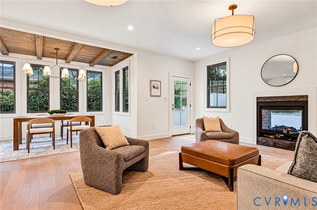 Living area featuring wood-finish flooring, exposed wood ceiling beams, a gas fireplace with a dark stone surround, drum-style pendant lighting, and multiple paneled windows