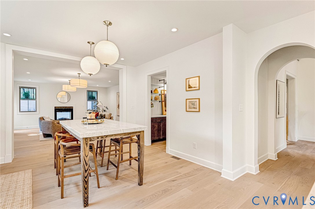 Open-concept interior featuring light wood-finish flooring, an arched entryway, recessed lighting, spherical pendant fixtures, and a stone-topped island