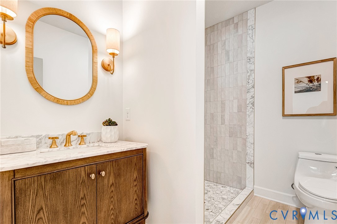 Wood-finish cabinetry featuring brass hardware, stone-top vanity with widespread faucet, oval mirror with textured frame, dual wall-mounted light fixtures, and walk-in shower with vertical tile surround