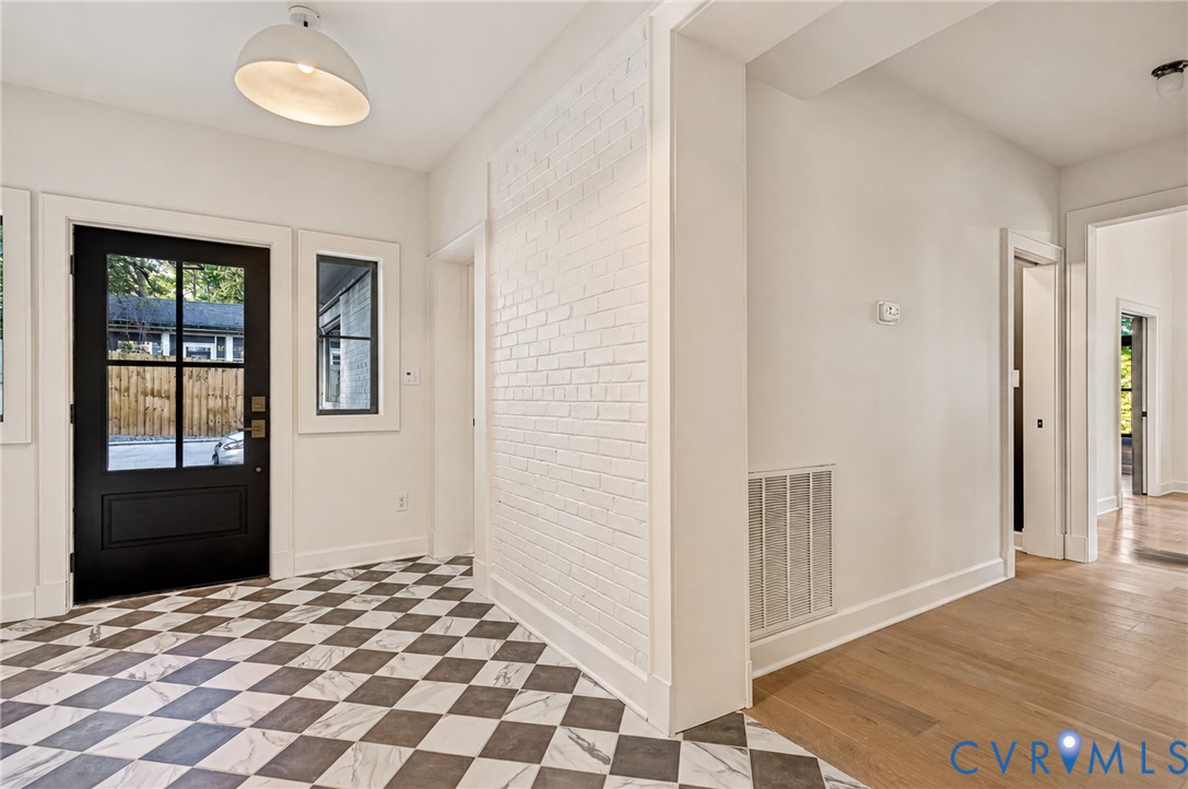 Entryway featuring black-framed multi-pane front door, checkerboard pattern tile flooring, wood-finish flooring, and white-painted brick accent wall with dome pendant lighting