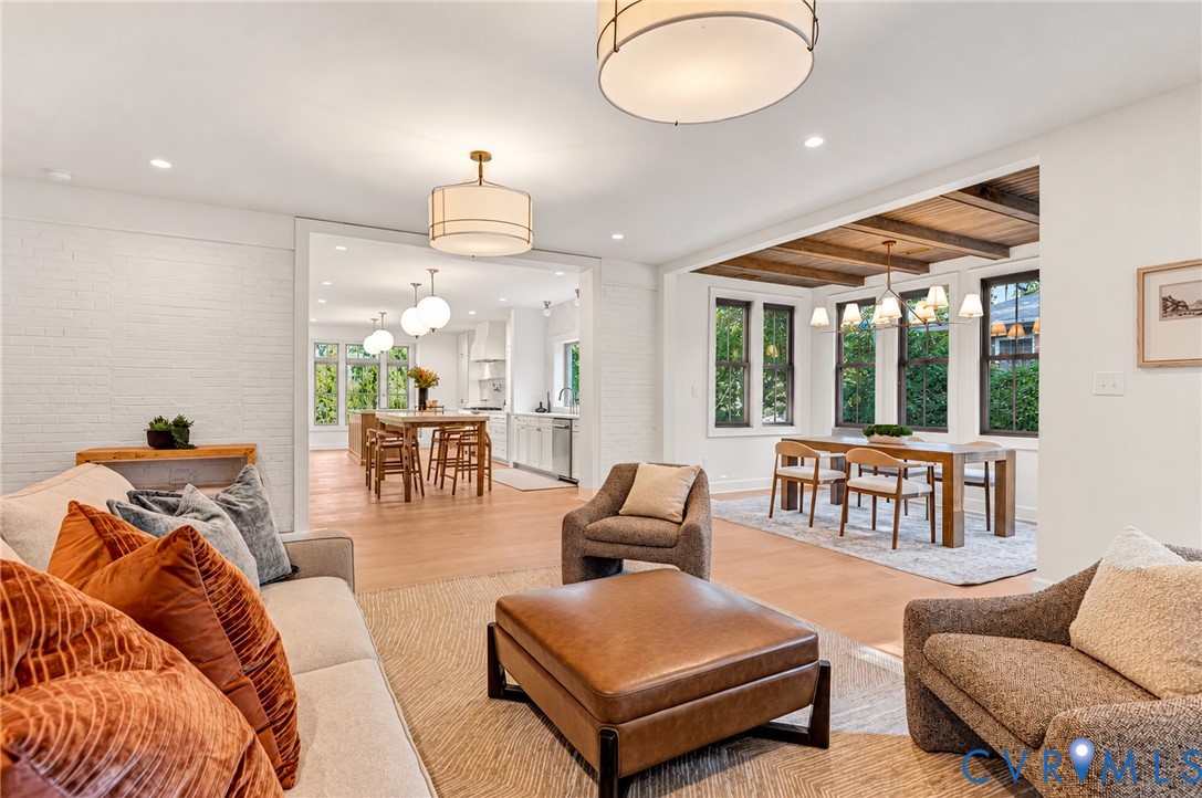 Open-concept area featuring white-painted brick walls, wood-finish flooring, recessed lighting, drum-style chandeliers, and exposed wooden ceiling beams