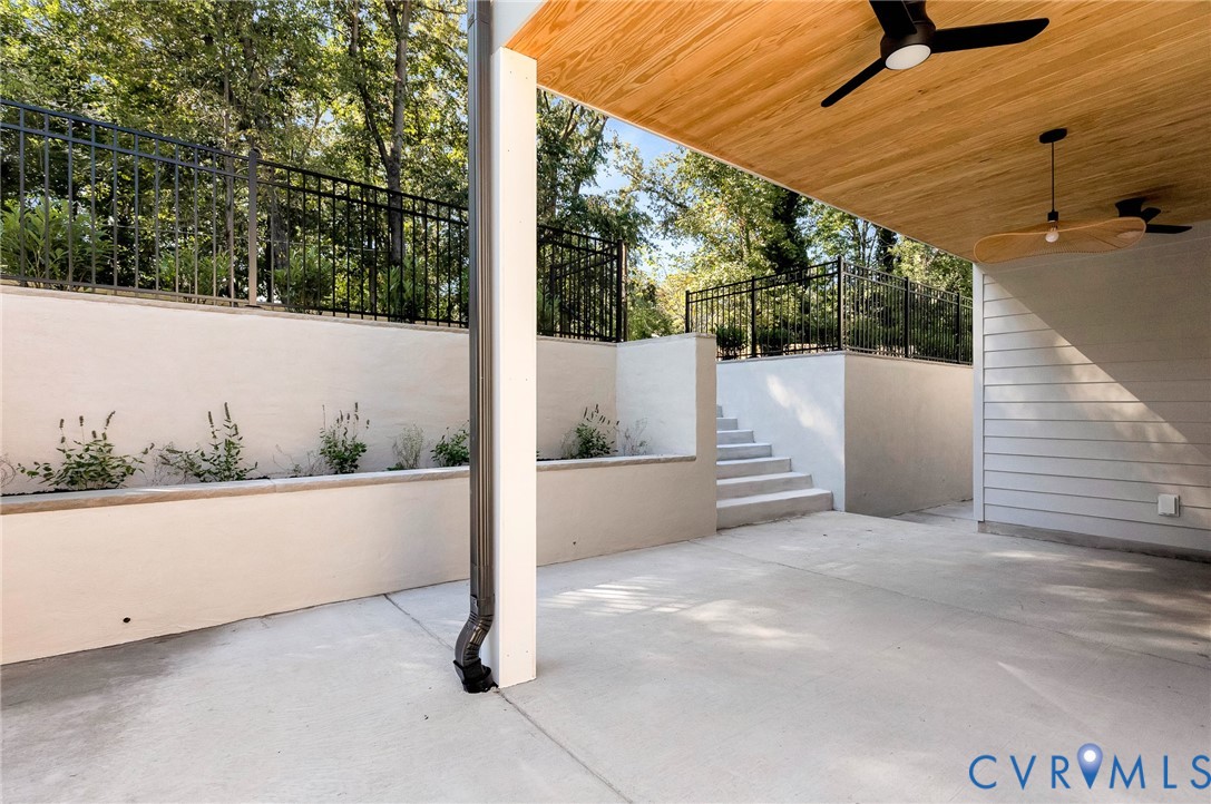 Covered patio with a wood-finished ceiling, integrated ceiling fans, concrete flooring, tiered retaining walls with metal fencing, and exterior stairs