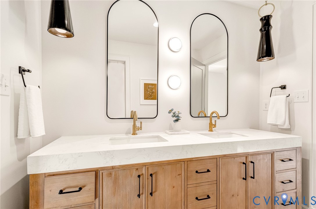 Double vanity featuring a stone-look countertop, integrated dual sinks, natural wood-finish cabinetry, arched mirrors with matte black frames, and brass-tone fixtures
