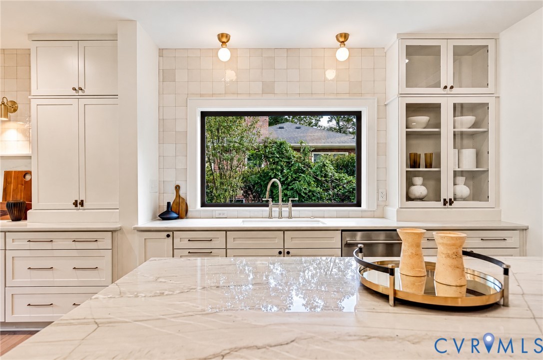 Kitchen featuring custom floor-to-ceiling cabinetry, stone countertops, square-tile backsplash, glass-front upper cabinets, and a wide window above the sink Kitchen featuring custom floor-to-ceiling cabinetry, stone countertops, square-tile backsplash, glass-front upper cabinets, and a wide window above the sink