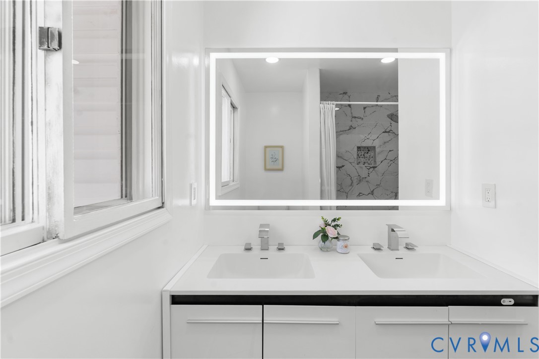 Dual undermount sinks with chrome faucets, illuminated mirror, and a window providing natural light