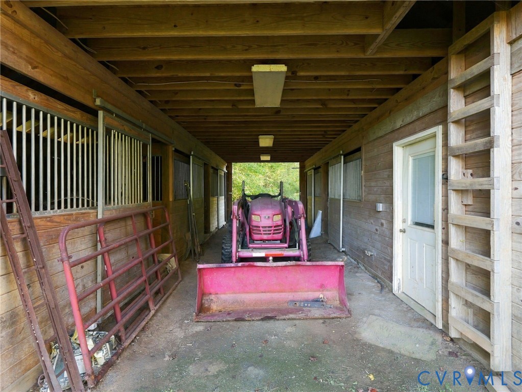 Covered stable area featuring wood-paneled walls, exposed wood ceiling joists, vertical metal stall partitions, and an integrated exterior door