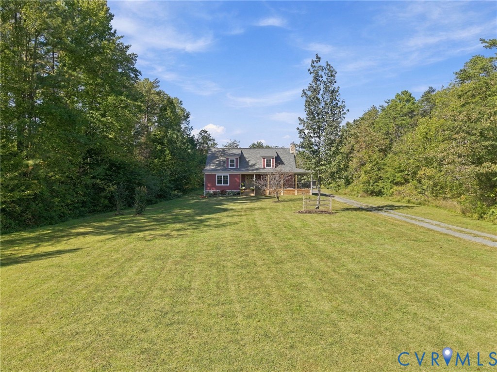 Cape Cod-style residence featuring a covered front porch, dormer windows, and a gravel driveway situated on a large, grass-covered lot surrounded by mature trees Cape Cod-style residence featuring a covered front porch, dormer windows, and a gravel driveway situated on a large, grass-covered lot surrounded by mature trees