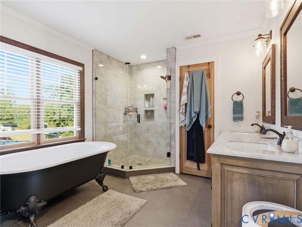 Freestanding clawfoot tub, dual-sink vanity with stone countertop, walk-in glass-enclosed shower featuring tile surround and built-in recessed shelving, and dark-toned cabinetry