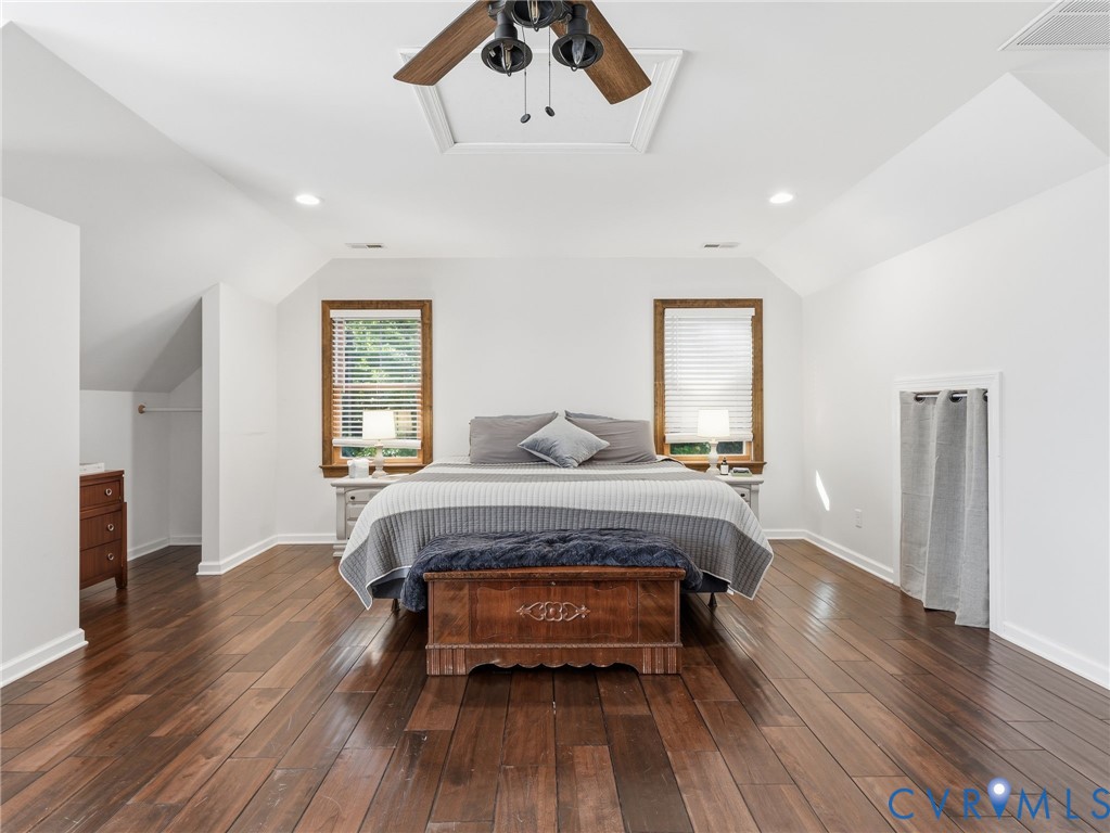Spacious room featuring wood-finish flooring, vaulted ceilings, two windows with wooden trim, and a ceiling fan with light fixture