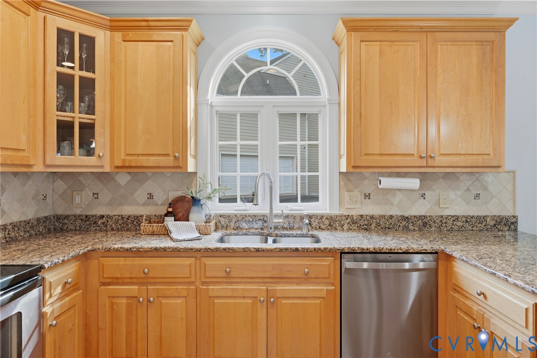 Kitchen featuring light wood cabinetry with glass-panel upper doors, stone-finish countertops, tile backsplash with decorative inserts, stainless steel double-basin sink with gooseneck faucet, and an arched window