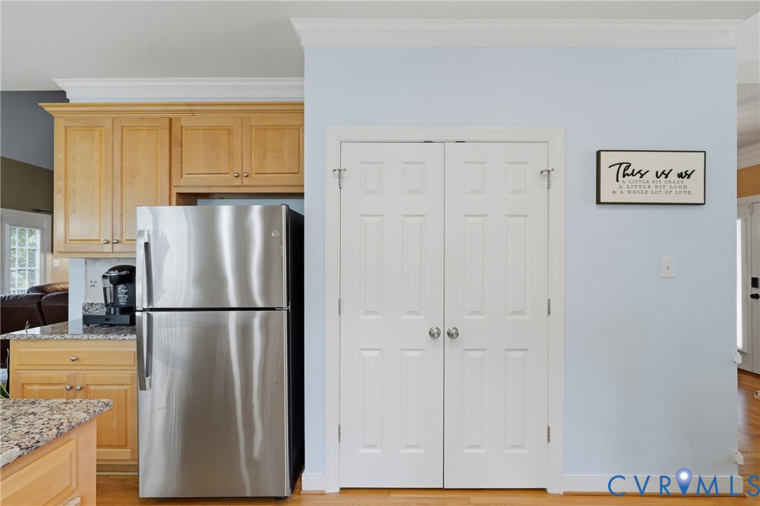 Kitchen area featuring light-toned wood cabinetry, stone countertops, a stainless steel refrigerator, and dual-panel closet doors with crown molding