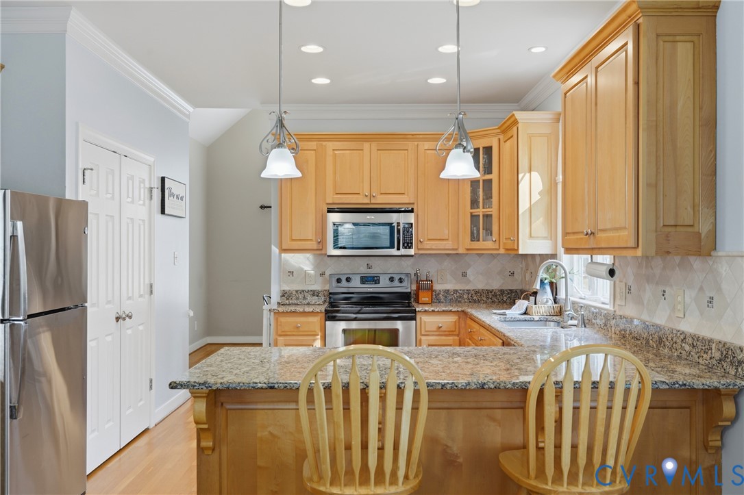 Kitchen featuring light wood cabinetry, stone countertops, stainless steel appliances, a diamond-pattern tile backsplash, and dual pendant lighting