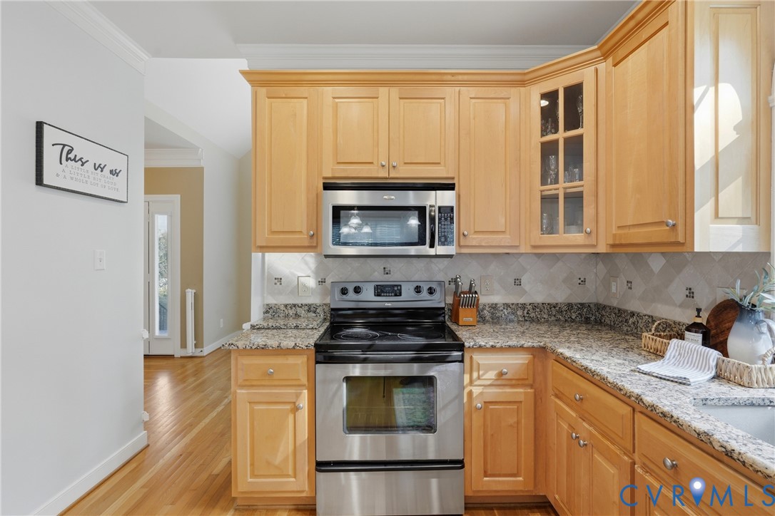 Kitchen featuring light wood cabinetry, stone countertops, stainless steel appliances, a glass-panel cabinet door, and chevron-patterned backsplash tile