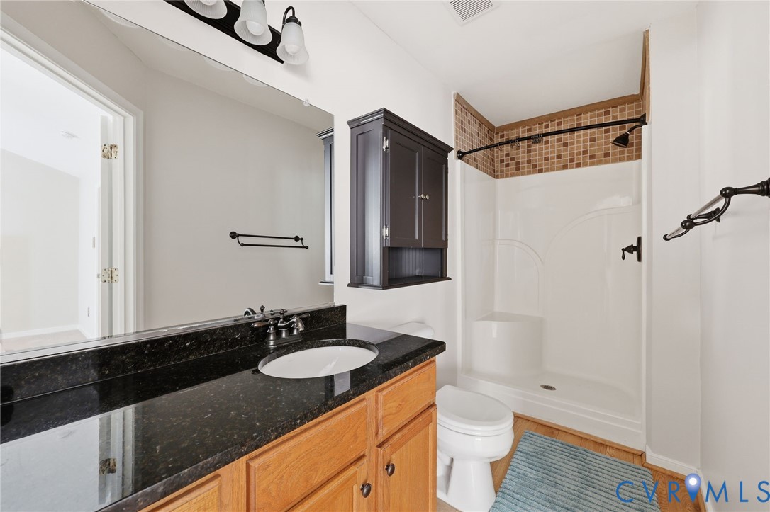 Dark stone-finish countertop with an integrated oval basin, natural wood-finish vanity cabinetry, wall-mounted dark cabinetry, and shower enclosure featuring tile-accented walls and built-in shelving