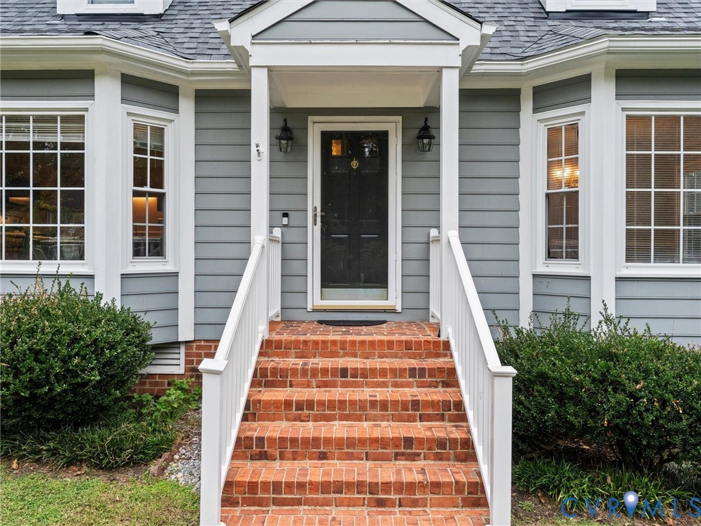 Exterior entrance featuring horizontal lap siding, multi-pane bay windows, a covered portico with white railings, and brick masonry steps Exterior entrance featuring horizontal lap siding, multi-pane bay windows, a covered portico with white railings, and brick masonry steps