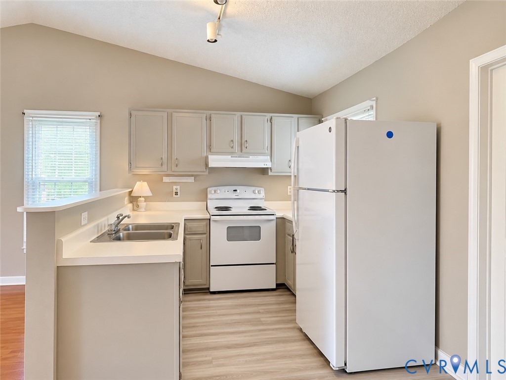 Kitchen featuring cabinetry with shaker-style doors, solid-surface countertops, dual-basin stainless steel sink, wood-finish flooring, and vaulted ceiling Kitchen featuring cabinetry with shaker-style doors, solid-surface countertops, dual-basin stainless steel sink, wood-finish flooring, and vaulted ceiling