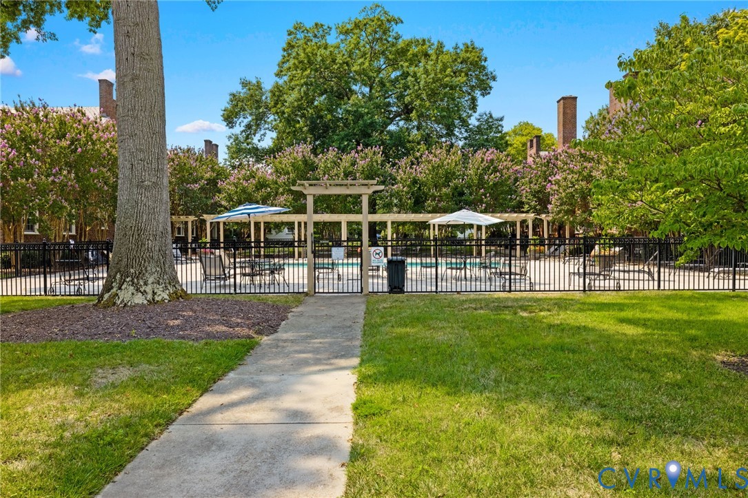 Swimming pool area featuring a metal perimeter fence, concrete walkway, and wooden trellis structure, complemented by manicured lawn and mature trees