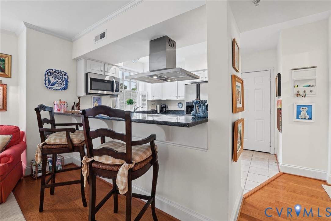 Kitchen featuring white cabinetry, dark stone-finish countertops, stainless steel wall-mounted range hood, built-in microwave, and breakfast bar seating area