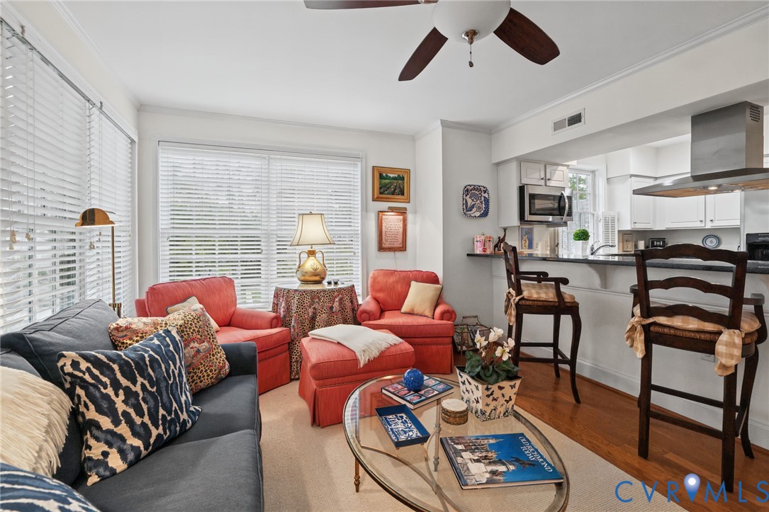 Open-concept living space featuring wood-finish flooring, a breakfast bar with dark countertops, white cabinetry, a ceiling fan, and large windows with horizontal blinds