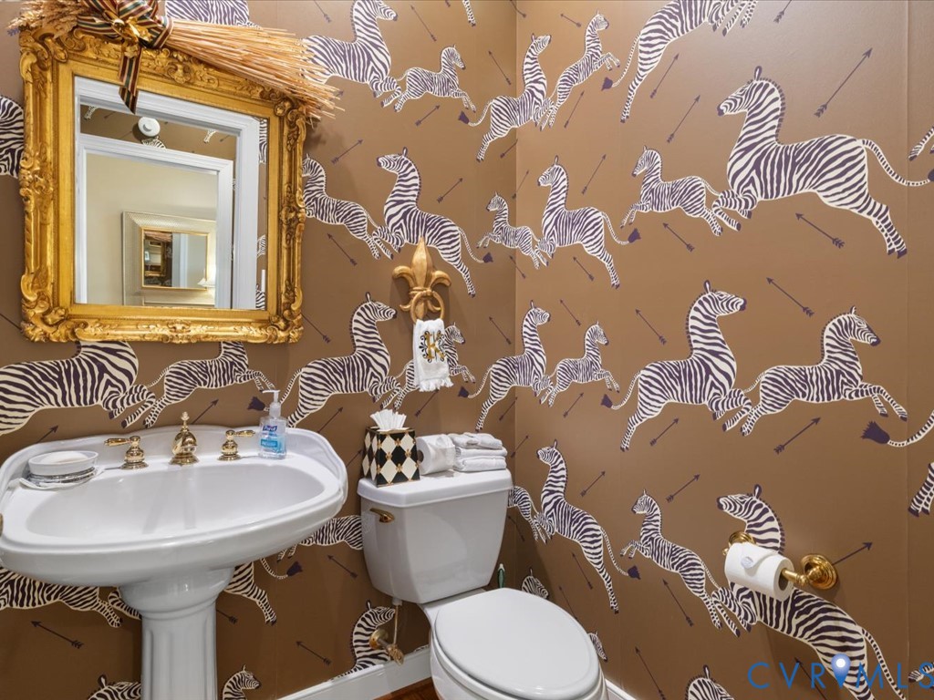 Pedestal sink with brass-finish faucet, toilet with matching brass-finish toilet paper holder, ornate gold-finish framed mirror, and distinctive patterned wallpaper