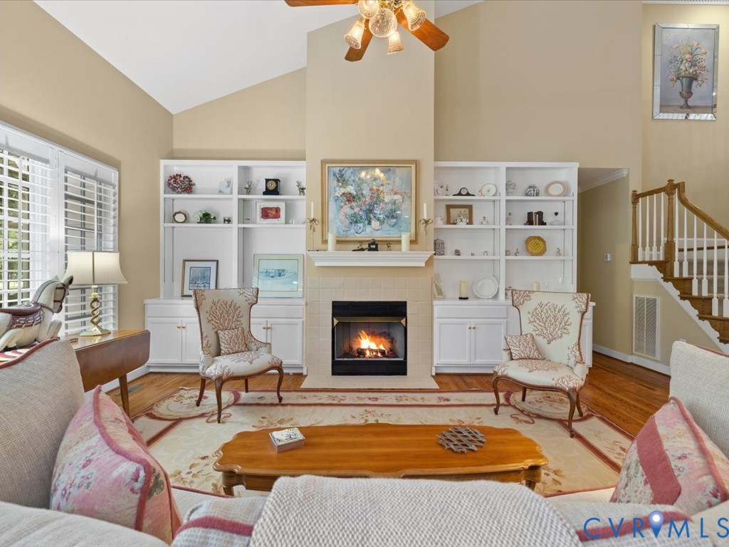 Vaulted ceiling with a ceiling fan light fixture, integrated built-in shelving, fireplace with a mantel, and wood flooring