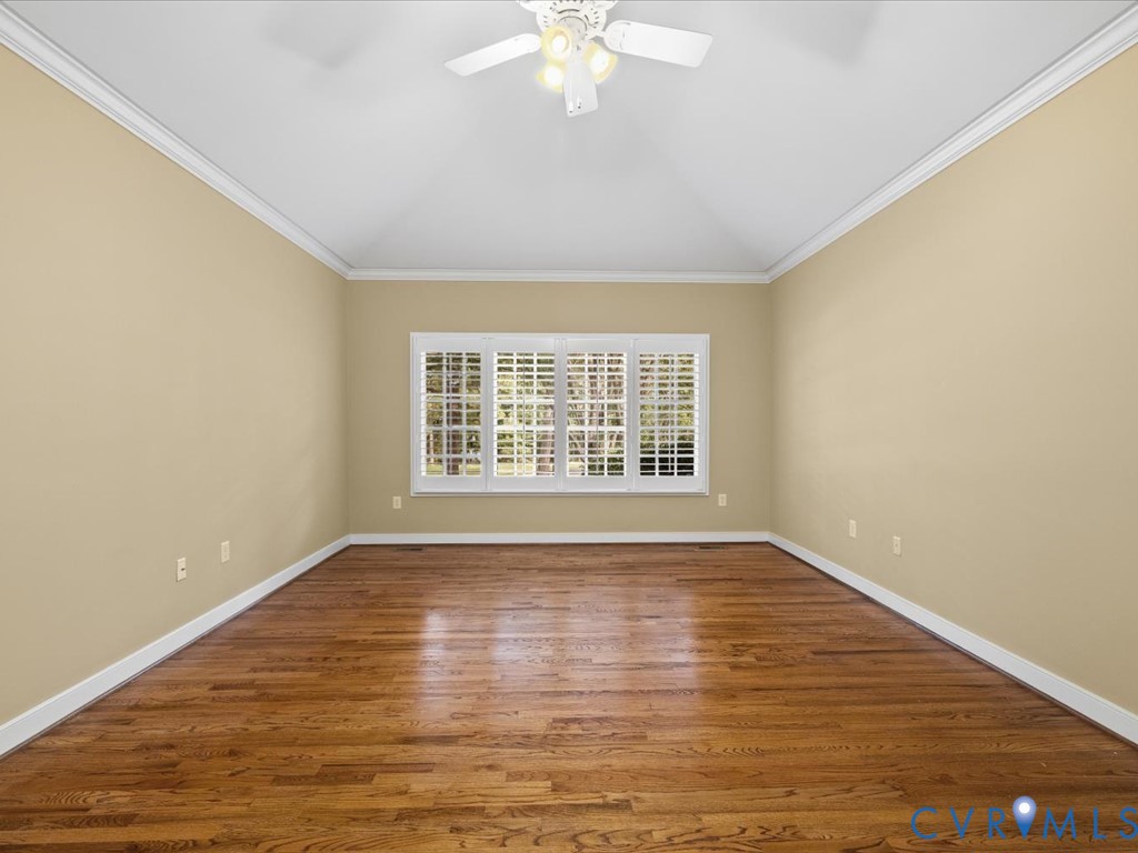 Vaulted ceiling with crown molding, ceiling fan with integrated lighting, wood-finish flooring, and a large window with white plantation shutters