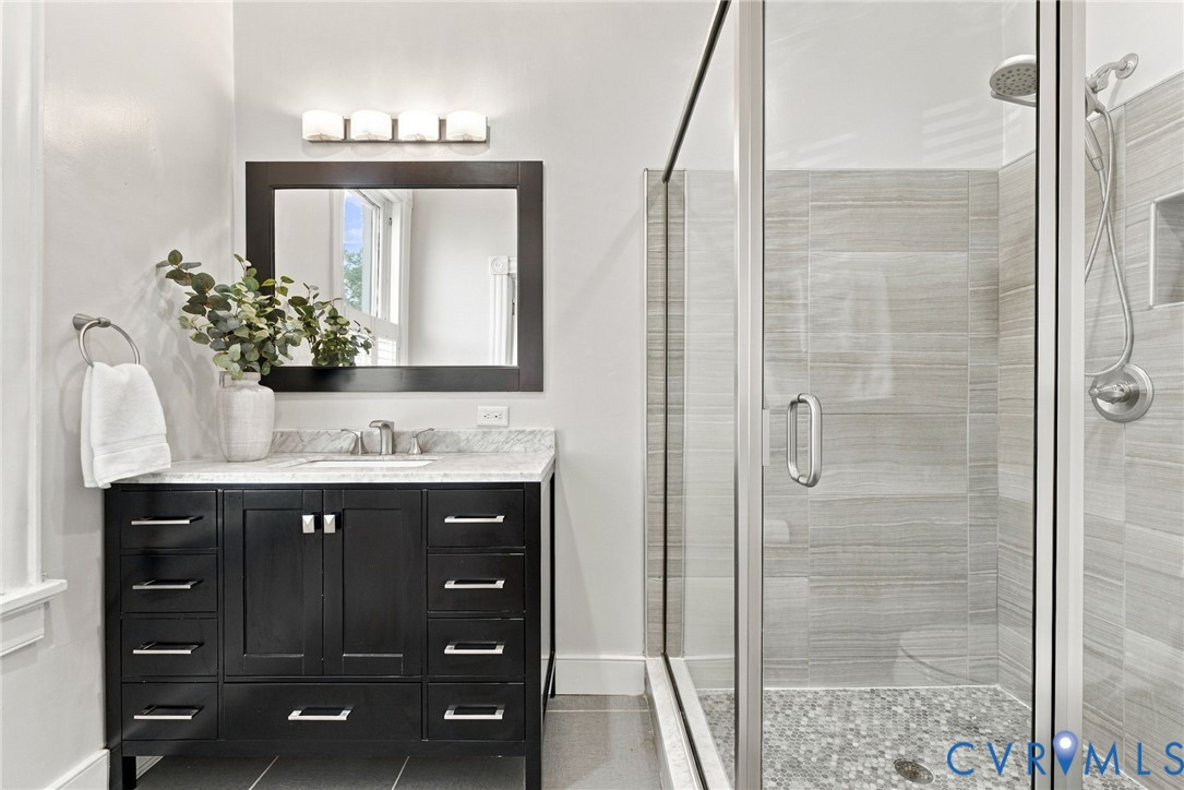 Dark wood-finish vanity with stone countertop, rectangular undermount sink, framed mirror with integrated lighting, and a glass-enclosed walk-in shower featuring horizontal wall tile and a stone-tiled basin
