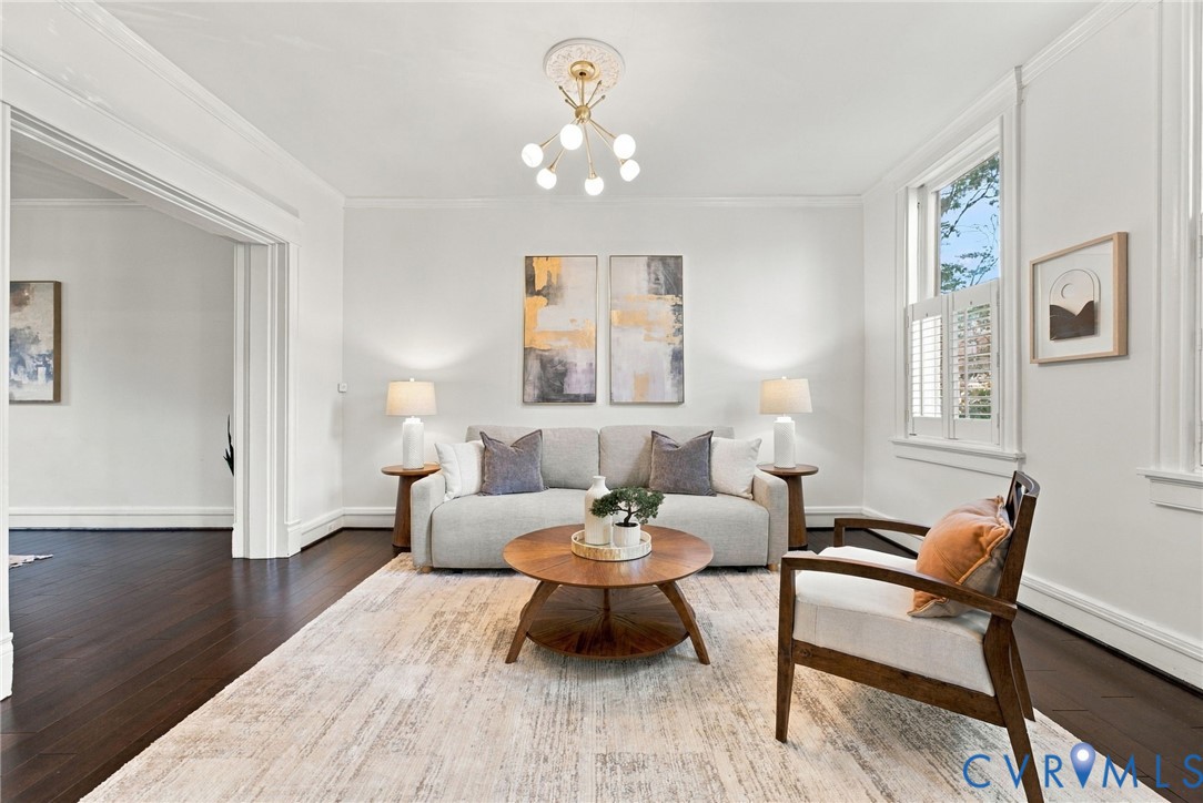 Living area featuring dark wood-finish flooring, crown molding, a central multi-arm light fixture with a decorative ceiling medallion, and plantation shutters