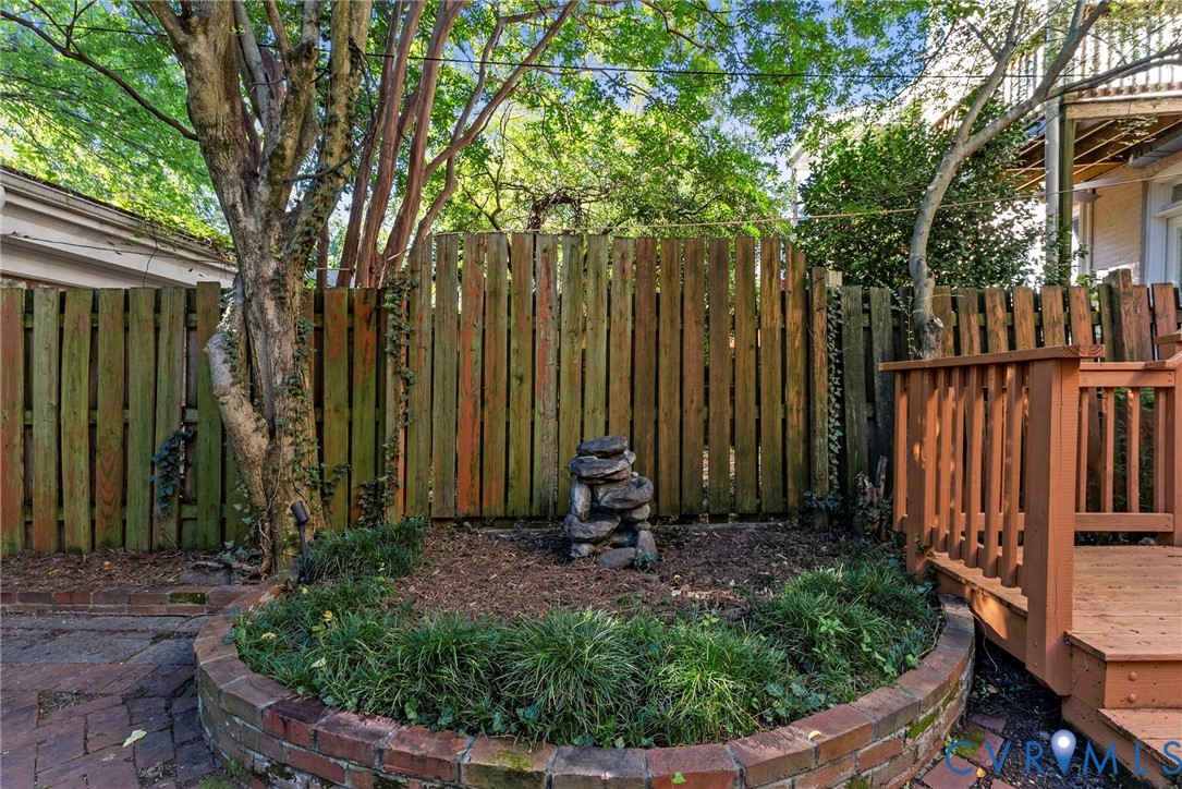 Rear yard featuring a wood-plank privacy fence, brick-bordered planter bed, wood-finish decking, and mature trees