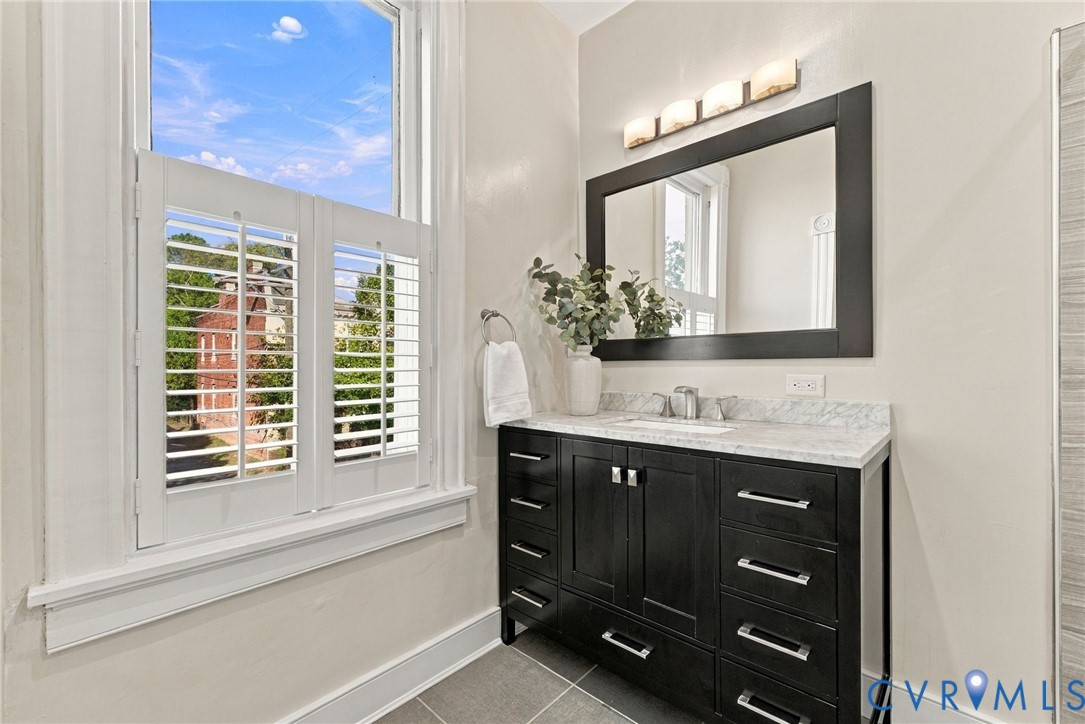 Dark wood vanity featuring a stone countertop with integrated sink, rectangular mirror with dark frame, four-light wall fixture, and white plantation shutters