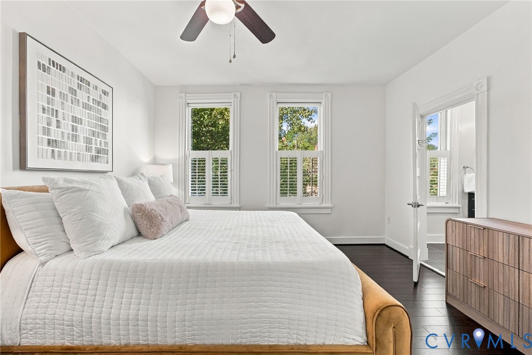 Bedroom featuring dark wood-finish flooring, two windows with white plantation shutters, and a ceiling-mounted fan with integrated light fixture