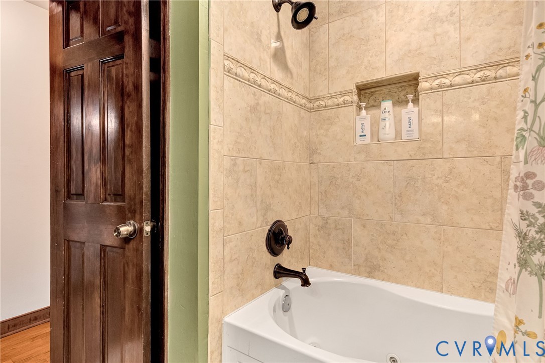 Soaking tub with a matte black faucet and shower head, surrounded by neutral-toned stone-finish wall tile, an integrated recessed soap niche, and a decorative tile border