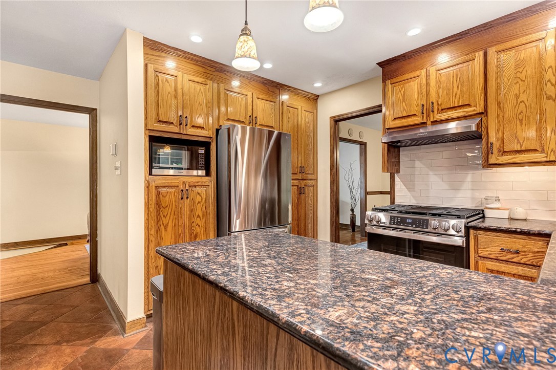 Kitchen featuring wood cabinetry, stone countertops, stainless steel appliances, a white tile backsplash, and overhead pendant lighting
