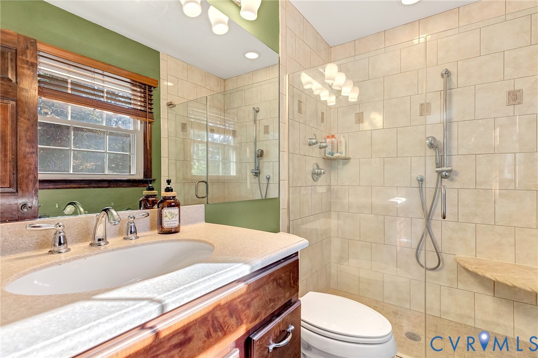 Bathroom featuring a wood-finish vanity with an integrated sink, polished chrome faucet, large frameless glass-enclosed shower with tile surround, built-in corner shower bench, and adjustable handheld shower head