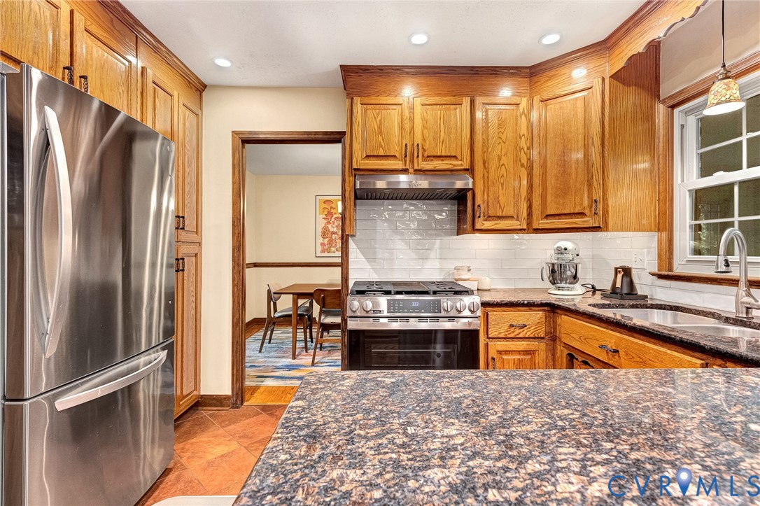 Kitchen featuring wood cabinetry, stone countertops, stainless steel refrigerator, gas range with integrated vent hood, and white subway tile backsplash