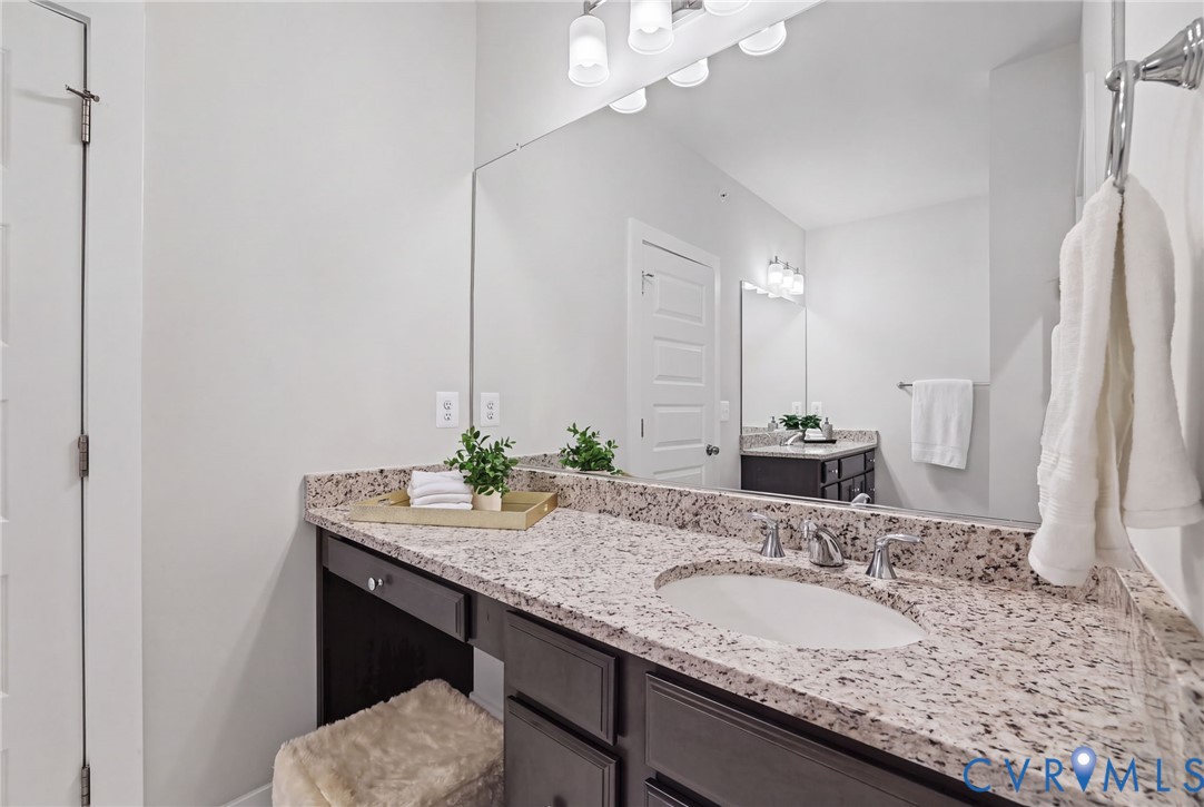 Dark-toned cabinetry featuring an integrated vanity, neutral stone-finish countertop, under-mount sink with dual-handle polished chrome faucet, and full-width wall-mounted mirror